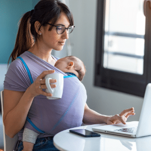 woman holding coffee working on laptop while holding sleeping baby in carrier