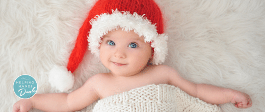Newborn baby with bright blue eyes looking at the viewer, wearing a Santa Claus hat. Baby is laying on a white, soft surface, with a knitted cream blanket over him.