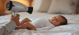 Newborn baby wearing white onesie is sleeping on bed while blow dryer is calming him being held by a parent.