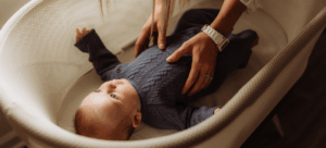 Newborn baby wearing a dark grey, knitted romper is being placed in a bassinet by their postpartum doula
