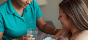 Helping Hands Doula wearing a teal coloured polo shirt brings a new mom a glass of water while she holds her newborn