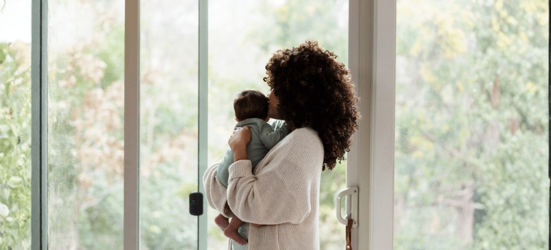 New mom holding and gently kissing her newborn by a sliding door with lots of soft light entering the room