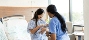 New mom wearing hospital gone is holding her newborn by the bed. Midwife with her hair tied back into a pony tail, looks over baby.