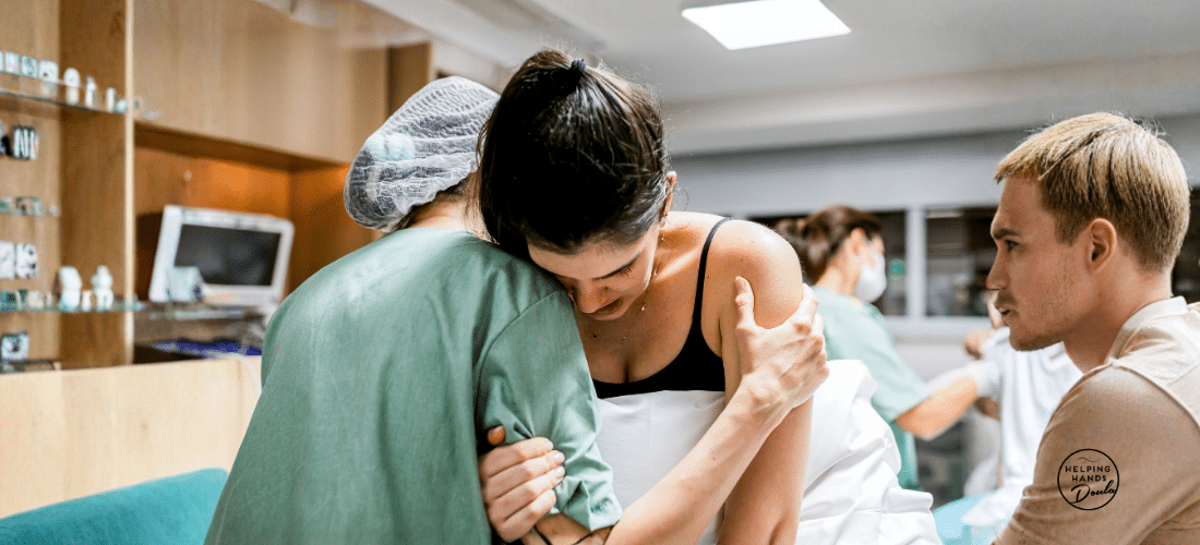 Calm childbirth scene of a birthing parent leaning into a doula, with her support parent beside her. In the distance a midwife and health care providers are ready for support.